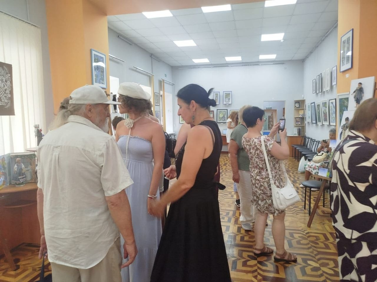Відвідувачі спілкуються, розглядають експонати та фотографують виставку авторських ляльок «КуклЬтура» у світлій залі Одеської бібліотеки. На передньому плані ліворуч розмовляють чоловік у кепці та жінка у блакитній сукні й береті на тлі інших гостей і картин на стінах.
