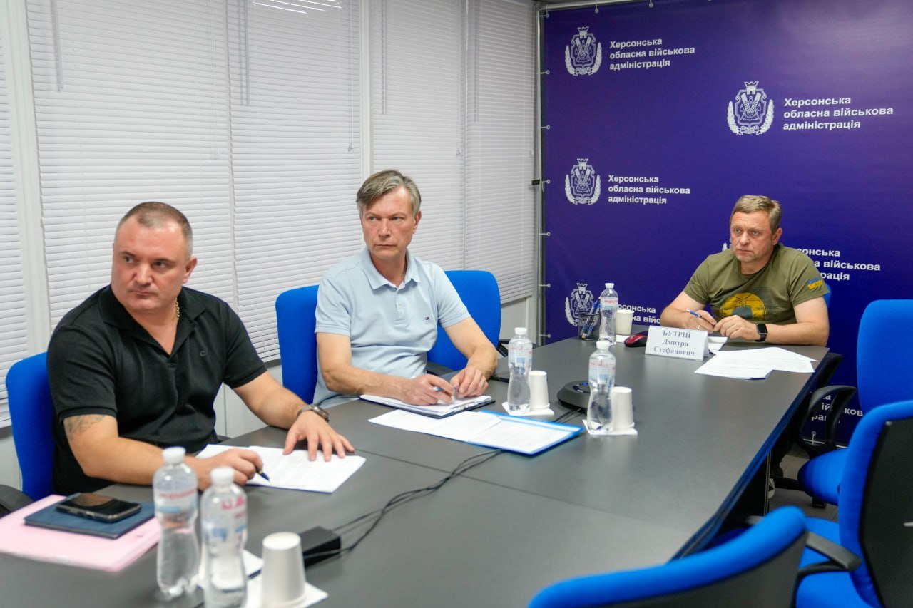 Перший заступник начальника Херсонської ОВА Дмитро Бутрій у зеленій футболці, заступник Херсонського міського голови Сергій Орєхов у світлій сорочці та член засідання в чорному поло сидять за сірим круглим столом із паперами та водою на тлі синього брендованого банера адміністрації.