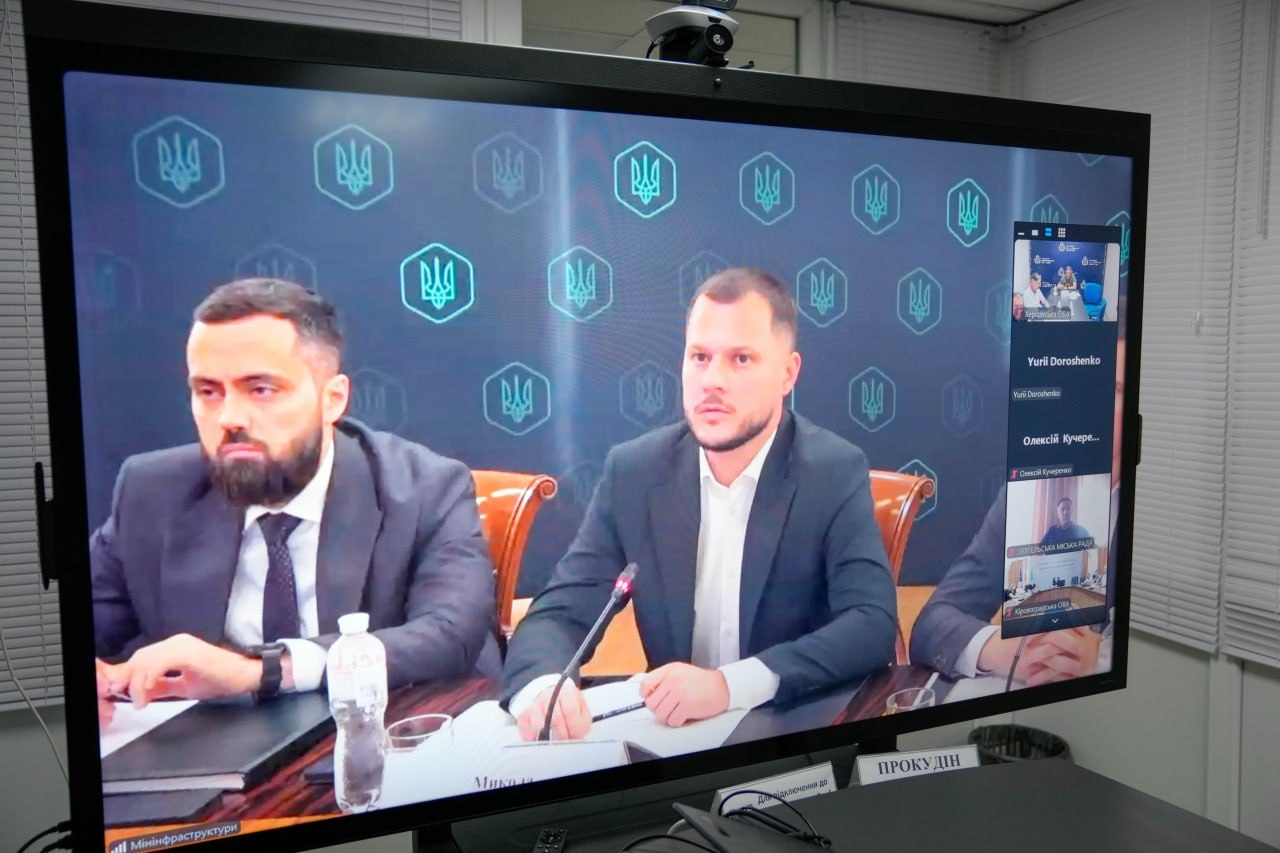 Екран телевізора під час онлайн-засідання Міністерства з відновлення, на якому видно учасників наради на тлі сірого брендованого банера з гербами України, а збоку відображаються вікна інших дистанційних підключень.