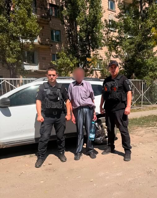 Поліцейські офіцери Дар'ївської громади у чорній формі супроводжують літнього чоловіка з розмитим обличчям під час евакуаційних заходів на тлі білого автомобіля та багатоповерхового будинку.