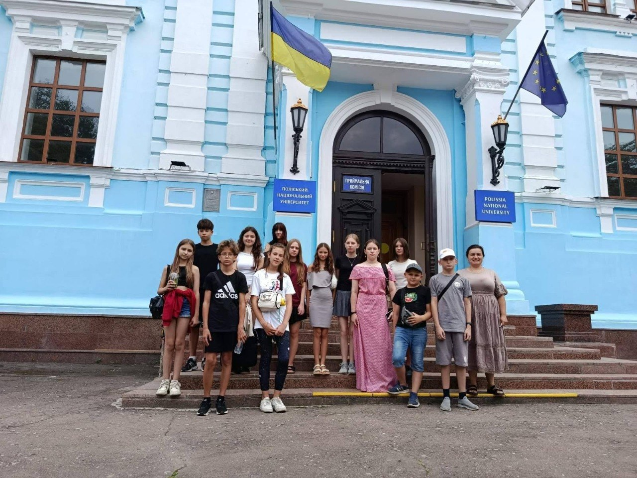 Група підлітків та молоді — переселенців із Херсонщини — разом із представниками координаційного центру «Вільні разом» міста Житомир стоять на сходах біля головного входу до Поліського національного університету в Житомирі під час профорієнтаційного заходу.