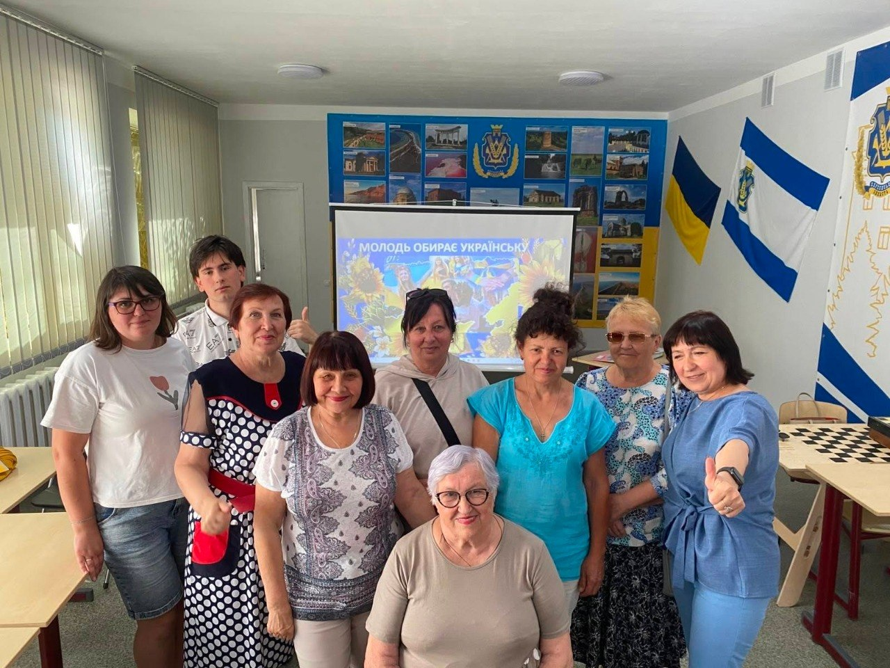 Група людей різного віку — переселенці з Херсонщини та представники координаційного центру «Вільні разом» міста Житомир — позують для спільного фото на тлі екрана з презентацією інтенсиву «Молодь обирає українську», прапора України та синьо-білого прапора Херсонської області на стіні.