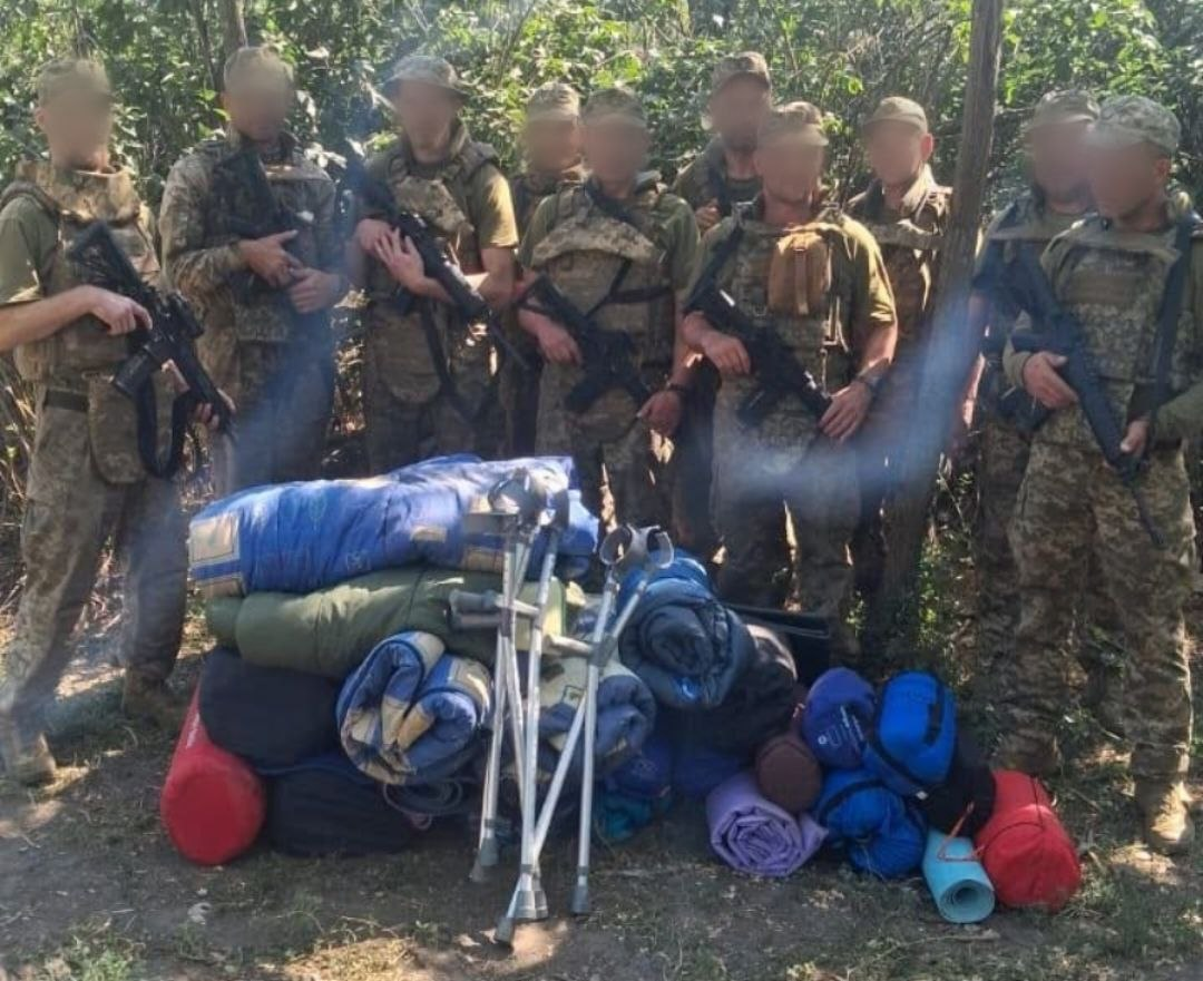 Група українських військовослужбовців у камуфляжній формі та зі зброєю стоїть серед дерев, позуючи біля переданої волонтерської допомоги від Верхньорогачицької СВА, БФ «Sunflower Seeds Ukraine» та Люка Хока, серед якої спальні мішки, каримати та медичні милиці.