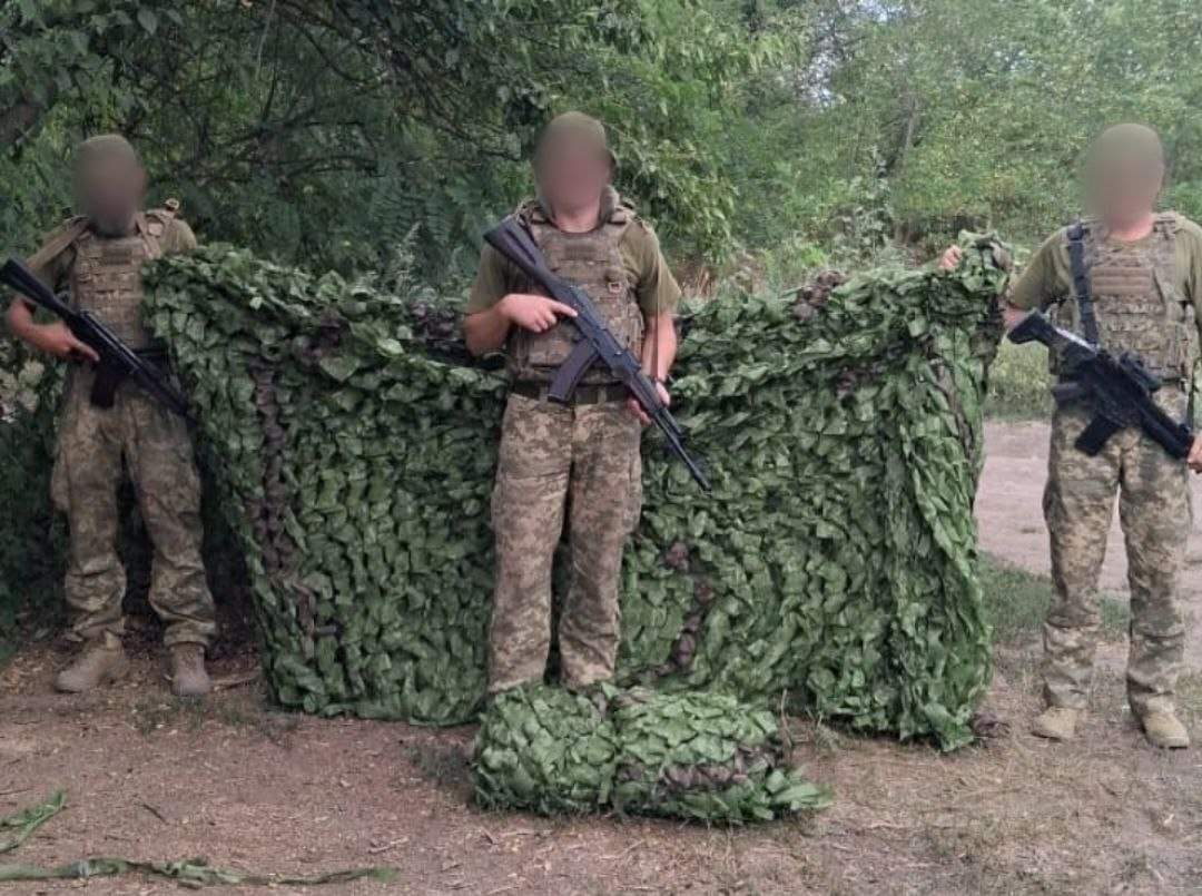 Три українські військовослужбовці з розмитими обличчями, у камуфляжній формі та зі зброєю, тримають розгорнуту велику зелену маскувальну сітку, отриману від Верхньорогачицької СВА та волонтерів, на тлі дерев.