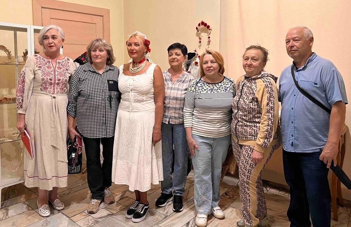 Групове фото переселенців із Херсонщини та організаторів на виставці артефактів Приазов’я в Івано-Франківському краєзнавчому музеї. Сім людей стоять у залі на тлі скляної вітрини з експонатами. Серед присутніх — майстриня в автентичному білому народному строї з восковим віночком на голові та жінка у вишитій сукні, інші гості вбрані у повсякденний одяг.