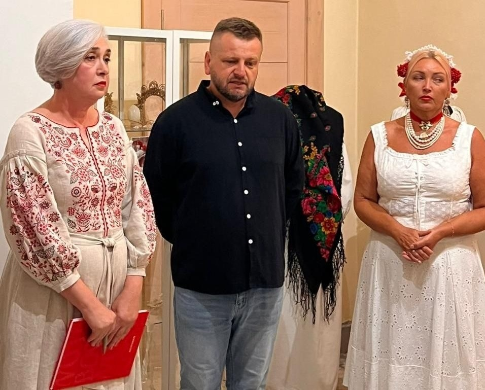 Організатори та майстриня під час презентації виставки артефактів Приазов’я в Івано-Франківську. Зліва направо стоять: жінка у вишитій сукні з червоною текою в руках, чоловік у чорній сорочці та джинсах, а також майстриня з лівобережжя Херсонщини в автентичному білому строї, традиційних намистах і восковому віночку з червоними квітами. На задньому плані видно скляну вітрину та старовинну українську хустку.