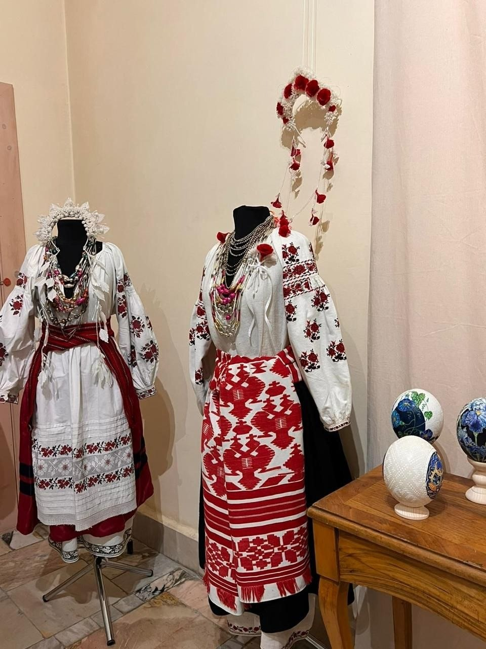 Експозиція автентичного українського вбрання та оберегів на виставці артефактів Приазов’я. На двох манекенах представлено традиційні жіночі строї Херсонської губернії: білі вишиті сорочки з червоно-чорними орнаментами, спідниці, фартухи-запаски та багатошарові коралові й металеві намиста. На голові одного з манекенів та на стіні вище розміщено традиційні воскові віночки з білими й червоними елементами. Справа на дерев'яному столику стоять три авторські писанки на підставках.