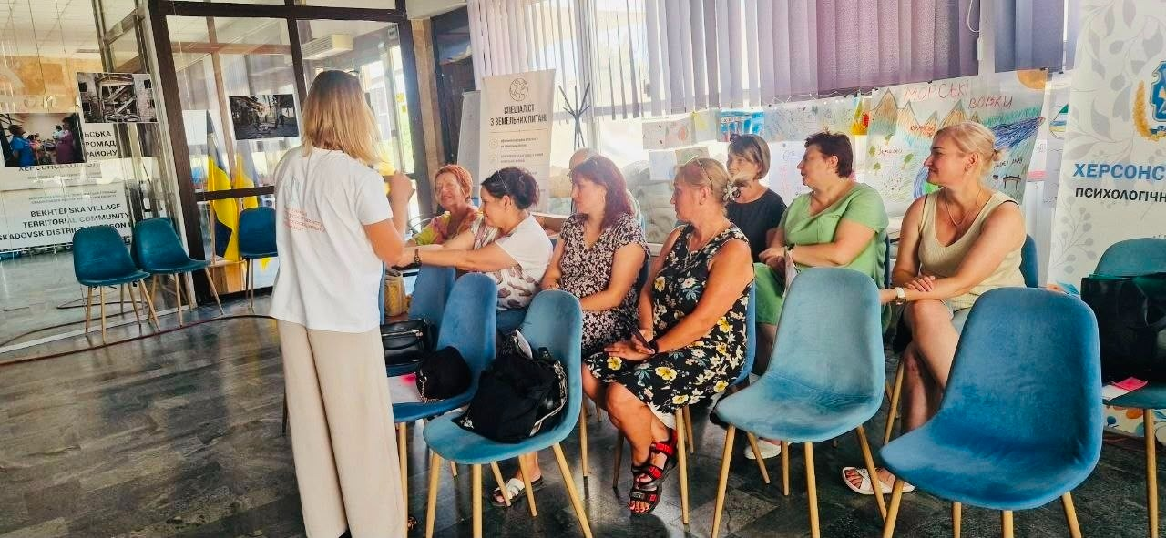 Зустріч переселенців із Херсонщини в миколаївському координаційному центрі «Вільні разом» на занятті з психосоціальної підтримки. Група учасниць сидить півколом на бірюзових стільцях та слухає психологиню, яка стоїть перед ними у білій футболці та світлих штанях. На стінах та інформаційних стендах позаду видно дитячі малюнки й банери з текстом.