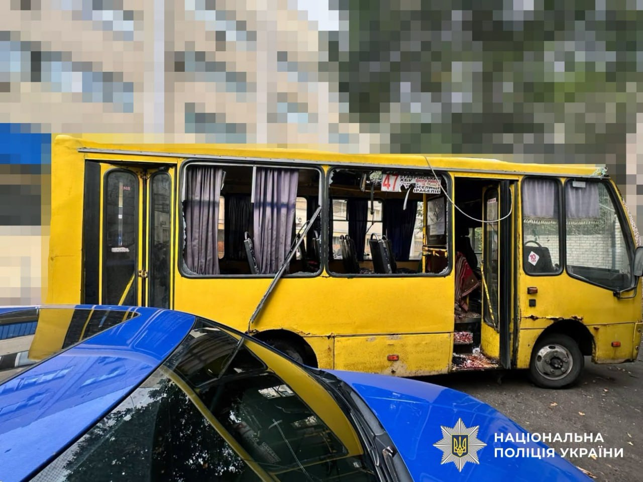 Жовтий міський автобус у Херсоні після атаки російського безпілотника, знятий з правого боку. На передньому плані видно синій капот автомобіля з логотипом Національної поліції України. У маршрутного таксі №47 повністю вибиті вікна пасажирського салону, пошкоджено лобове скло та відчинено передні двері, біля яких видно уламки.