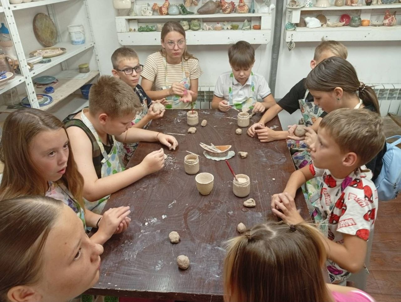 Майстер-клас із ліплення з глини в Одесі для дітей-переселенців із Херсонщини, вид згори. Група дітей у захисних фартухах сидить навколо великого коричневого столу, на якому розкладені шматочки глини, глиняні заготовки та баночки з водою чи шлікером. На чолі столу сидить молода дівчина-наставниця в окулярах, яка пояснює завдання. На задньому плані розташовані полиці з різноманітними готовими керамічними виробами та фігурками.