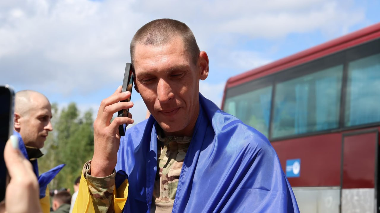 Звільнений із російського полону український захисник розмовляє по мобільному телефону одразу після обміну. Чоловік у камуфляжній формі загорнутий у синьо-жовтий прапор України, його очі прикриті від емоцій. На задньому плані видно червоний пасажирський автобус, іншого військового із поголеною головою та хмарне небо.