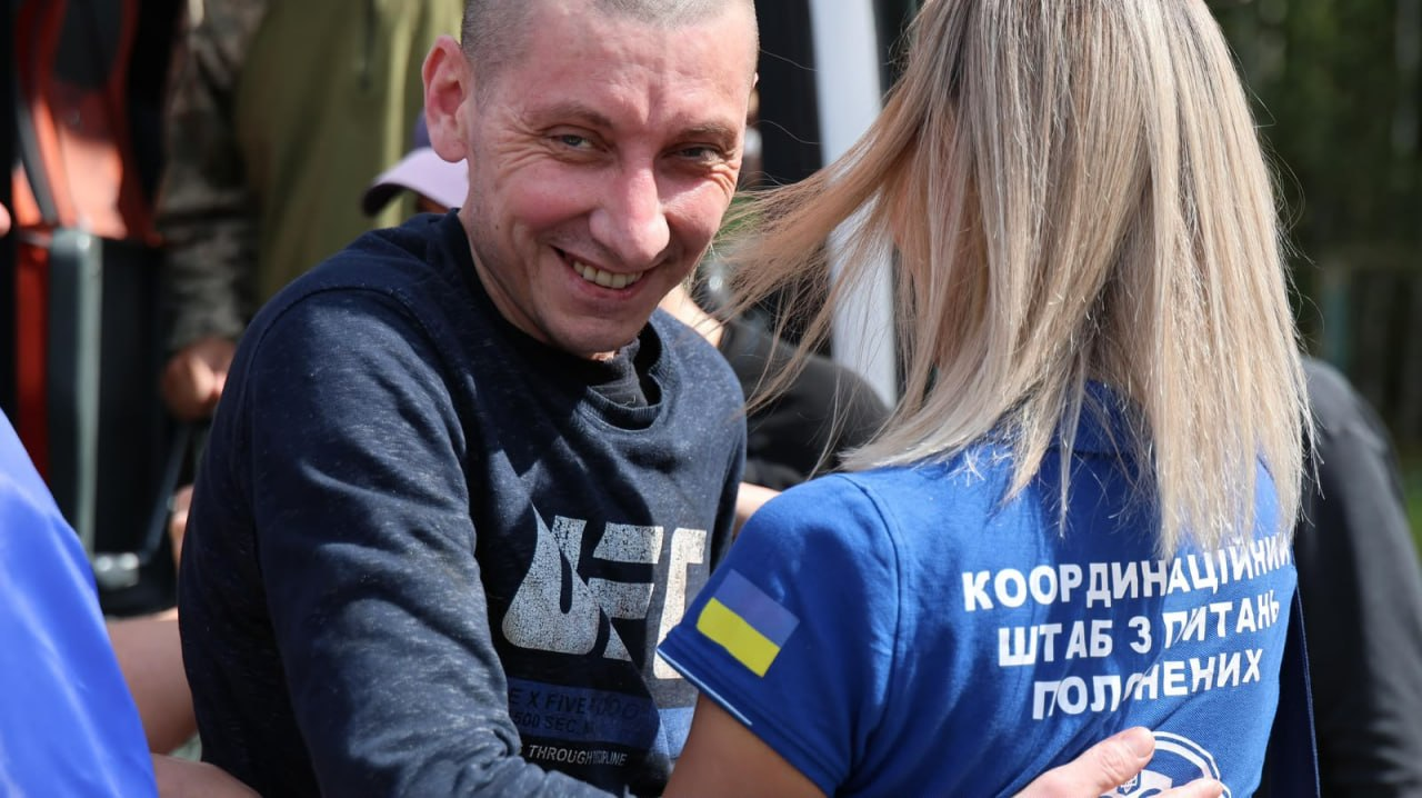 Усміхнений український захисник одразу після звільнення з російського полону обіймає представницю Координаційного штабу. Чоловік одягнений у темну кофту з принтом. Дівчина стоїть спиною до камери в синій футболці-поло з прапором України на рукаві та великим білим написом на спині: «Координаційний штаб з питань... полонених».
