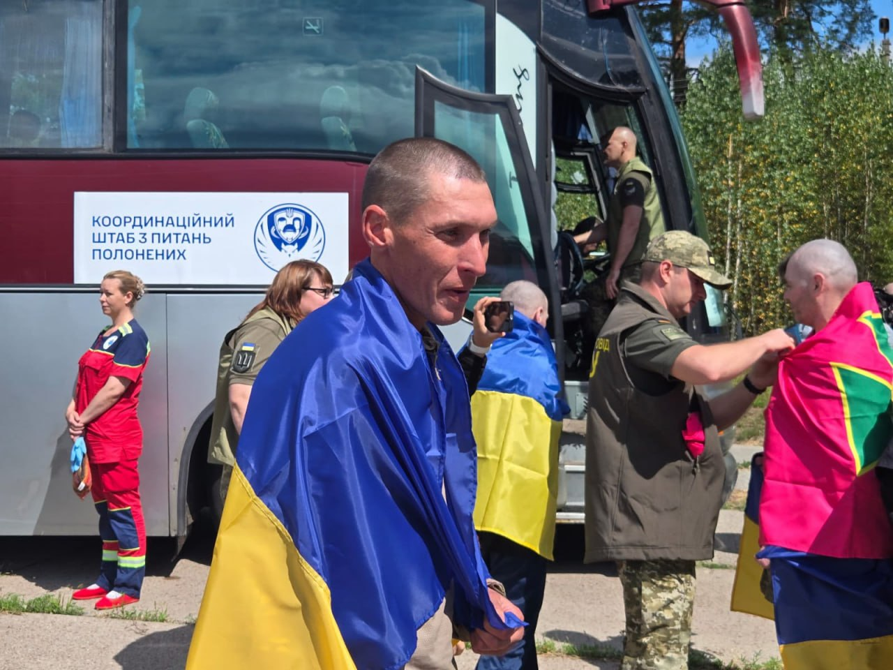Звільнені українські воїни біля автобуса під час обміну полоненими. На передньому плані — захисник у камуфляжній формі, загорнутий у синьо-жовтий прапор України. На борту пасажирського автобуса розміщено емблему та напис «Координаційний штаб з питань полонених». Поруч перебувають представники військових відомств, медики у червоній формі та інші повернуті з полону бійці.