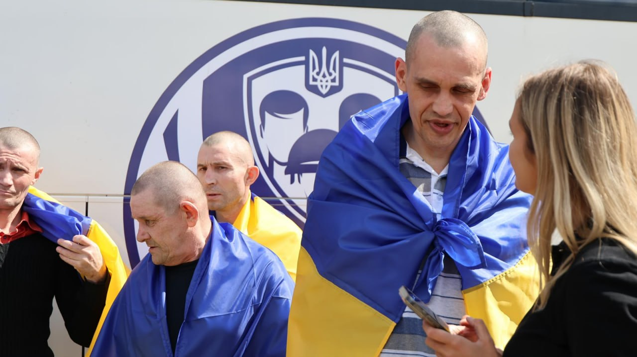 Звільнені з російського полону українські захисники під час обміну. На передньому плані — високий худорлявий військовослужбовець із поголеною головою, загорнутий у синьо-жовтий прапор України, який розмовляє з білявою дівчиною-представницею Координаційного штабу. Позаду них стоять ще троє визволених воїнів із прапорами на плечах. На білому борті автобуса нанесено великий круглий логотип Координаційного штабу з питань поводження з військовополоненими.