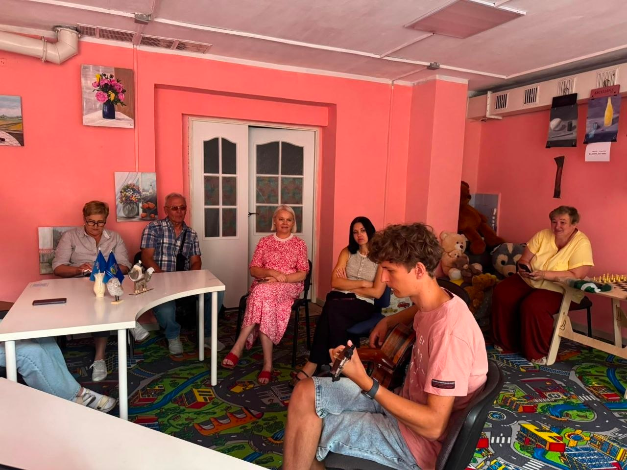 Творча зустріч та психологічна розвантаження для ВПО з Херсонщини у львівському хабі «Вільні разом». На фото зафіксовано момент музичного виступу: молодий виконавець із гітарою сидить у центрі кімнати перед аудиторією з переселенців, які уважно слухають музику в облаштованому для відпочинку приміщенні з рожевим інтер'єром.