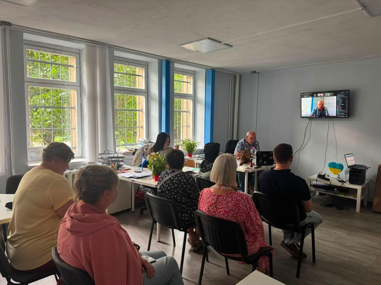 На світлині зафіксовано інформаційно-просвітницький захід: у світлому кабінеті з великими вікнами та сіро-блакитними стінами сидять слухачі — внутрішньо переміщені особи та працівники хабу. Їхня увага спрямована на великий настінний екран, на якому через відеозв'язок транслюється виступ лікаря-ендоскопіста та хірурга першої категорії Володимира Тамазликаря. На столі під телевізором облаштовано ноутбук, колонку та патріотичний декор у синьо-жовтих кольорах. Учасники зустрічі уважно слухають медичного фахівця, який розповідає про профілактику онкологічних захворювань шлунково-кишкового тракту.