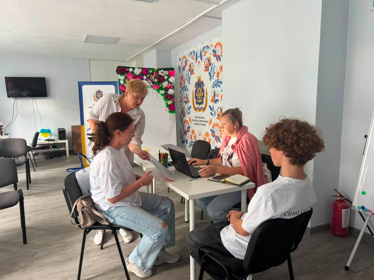 Індивідуальний прийом та практична допомога молоді з Херсонщини щодо роботи з онлайн-системою «Електронний суд» у львівському хабі «Вільні разом». На світлині зафіксовано робочий процес у центрі: за столом із ноутбуком співробітниця хабу консультує молодих переселенців, роз'яснюючи їм алгоритми дистанційного подання позовів, отримання судових документів та відстеження розгляду справ. Інша представниця організації допомагає дівчині з опрацюванням паперів, поки хлопець на передньому плані чекає на завершення консультації. Прийом проходить на тлі банера Херсонської ОДА та стіни з великим гербом України в традиційному стилі розпису.