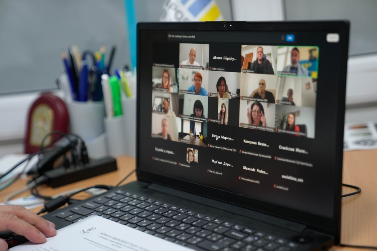 Засідання Комітету Верховної Ради України з питань гуманітарної та інформаційної політики в режимі відеоконференції.На світлині крупним планом зображено екран відкритого чорного ноутбука, на якому триває онлайн-нарада у форматі Zoom. На моніторі видно вікна з відеотрансляціями та іменами учасників засідання, серед яких посадовці та представники профільних служб. На передньому плані ліворуч рука учасника тримає паперовий документ із офіційним бланком Комітету Верховної Ради, а на задньому плані помітно робоче приладдя на офісному столі.