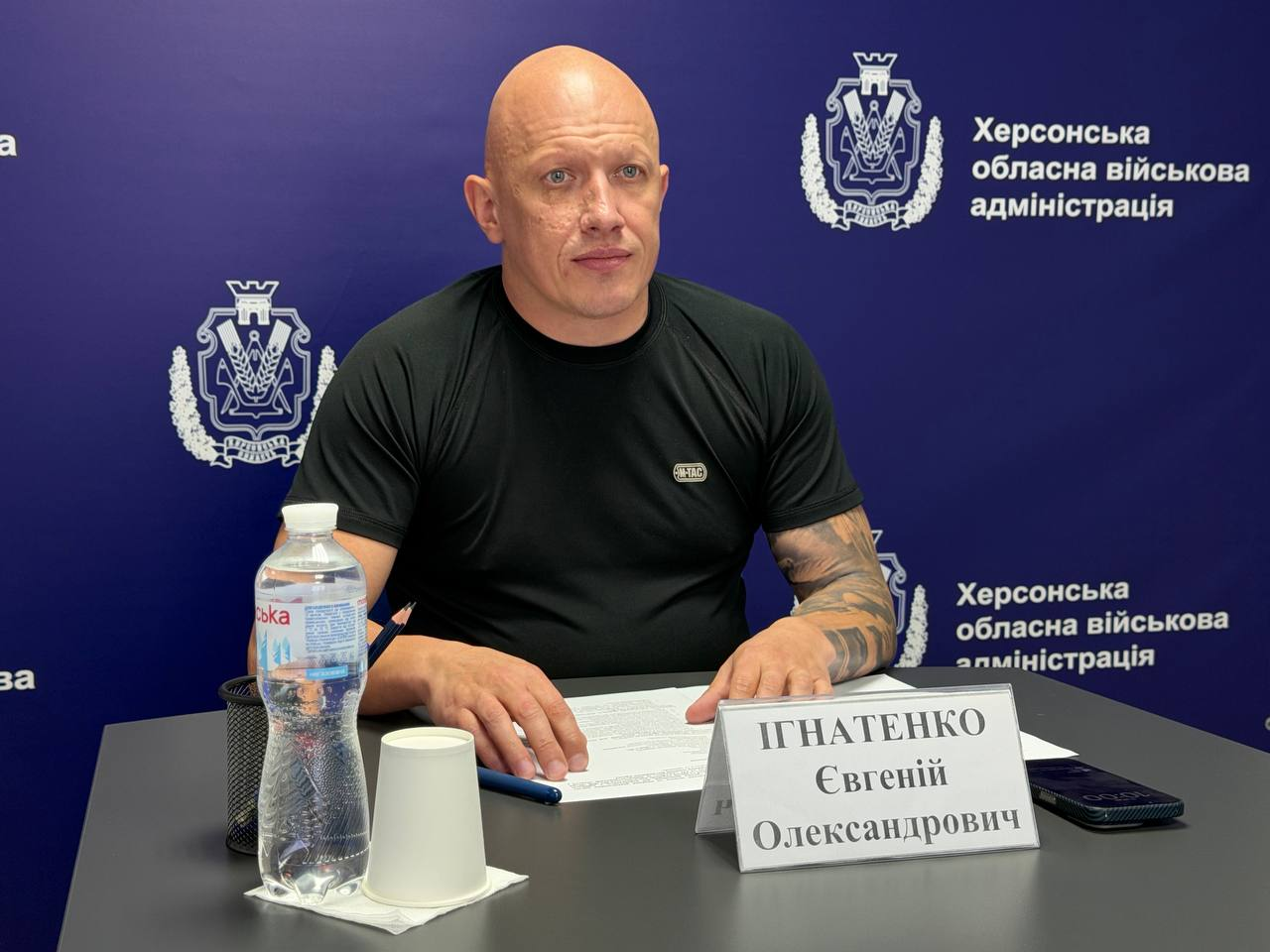 Заступник начальника Херсонської ОВА Євгеній Ігнатенко під час наради з представниками БФ «Вітри змін» щодо роботи мобільних бригад психосоціальної допомоги на Херсонщині. Чоловік сидить за столом на тлі синього банера з офіційними логотипами адміністрації.