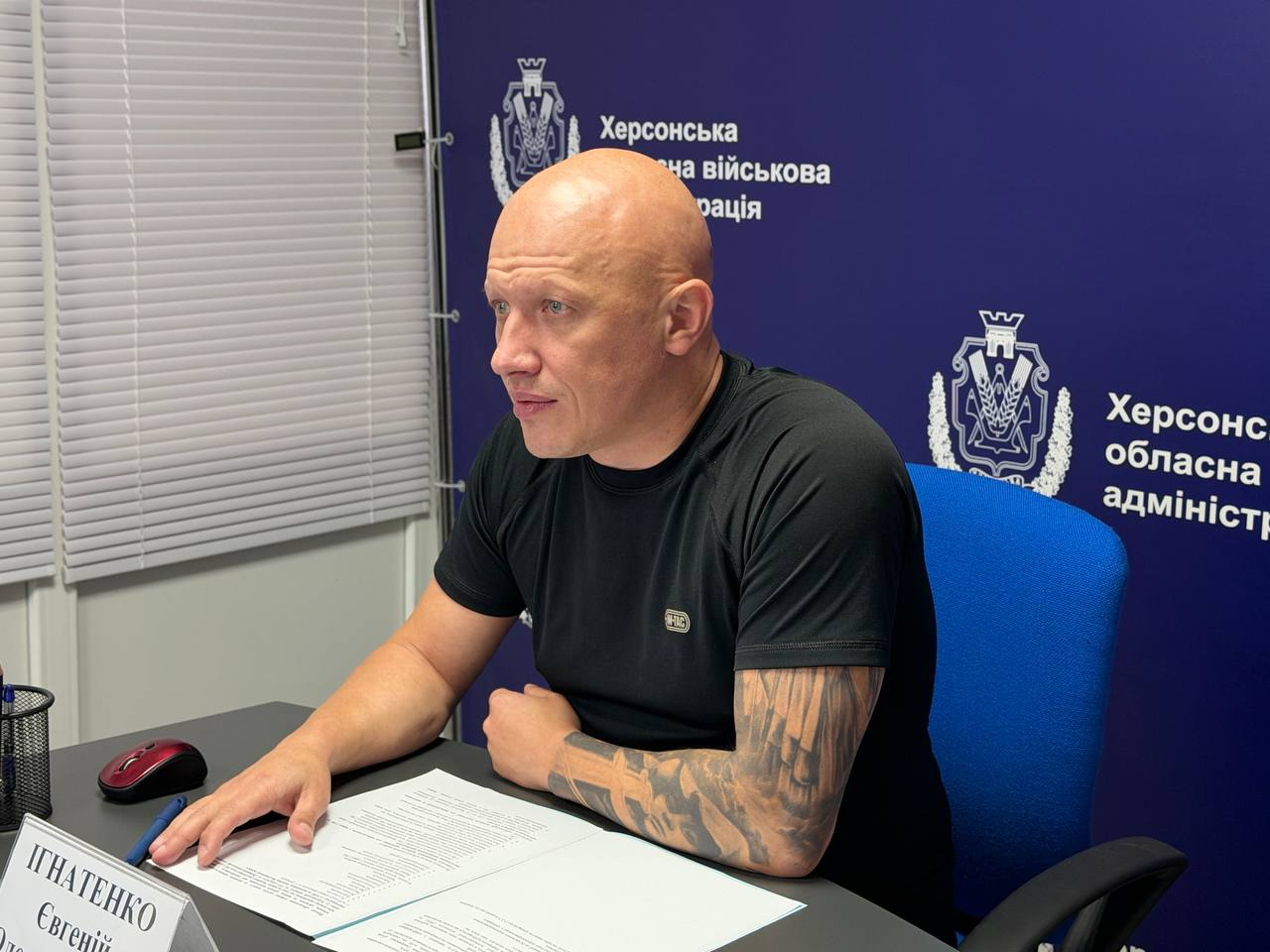 Заступник начальника Херсонської ОВА Євгеній Ігнатенко сидить у профілі за столом під час наради з БФ «Вітри змін» щодо психосоціальної допомоги. Чоловік дивиться ліворуч, перед ним лежать робочі документи та табличка з ім'ям, а на тлі видно синій банер із гербом та логотипом Херсонської обласної військової адміністрації.