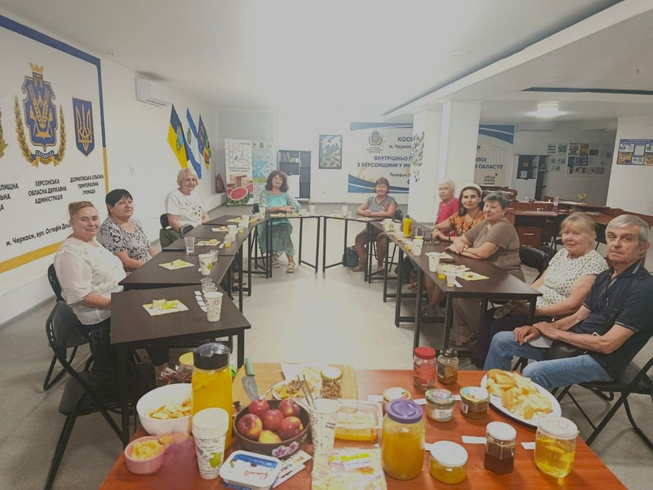 Загальний план заходу до Дня пасічника для переселенців з Херсонщини в координаційному центрі «Вільні разом» у Черкасах. Учасники сидять за столами, розставленими півколом у залі з прапорами та офіційними банерами Херсонської ОДА й Долматівської громади. На передньому плані стоїть стіл із дегустаційними частуваннями: банками меду, тарілками з яблуками, печивом та нарізаним хлібом.