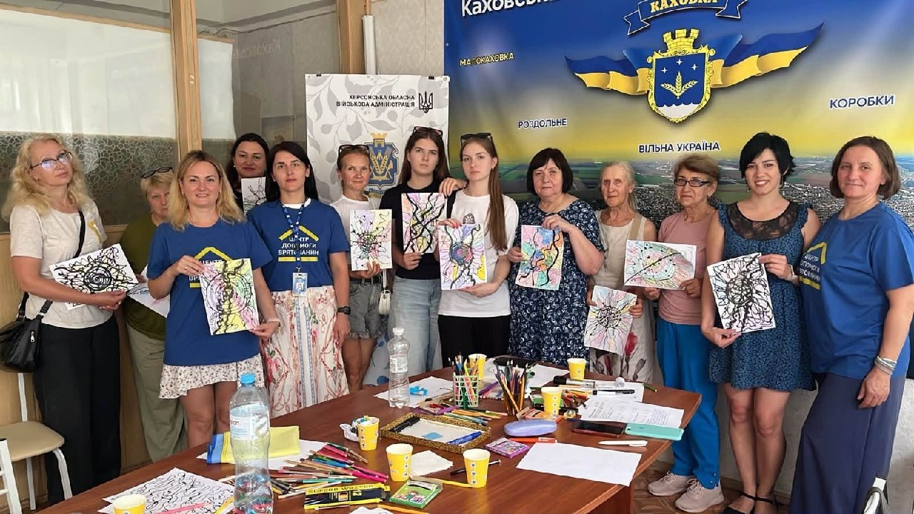 Групове фото учасниць заняття з нейрографіки для переселенок із Херсонщини в координаційному центрі «Вільні разом» у місті Києві. Жінки різного віку стоять в один ряд за великим робочим столом, демонструючи свої готові абстрактні малюнки з лініями та колами. На столі перед ними розкладено кольорові олівці, фломастери, склянки та пляшки з водою, а на тлі розміщено офіційний білий банер Херсонської ОВА та великий синьо-жовтий банер Каховки з гербом міста.