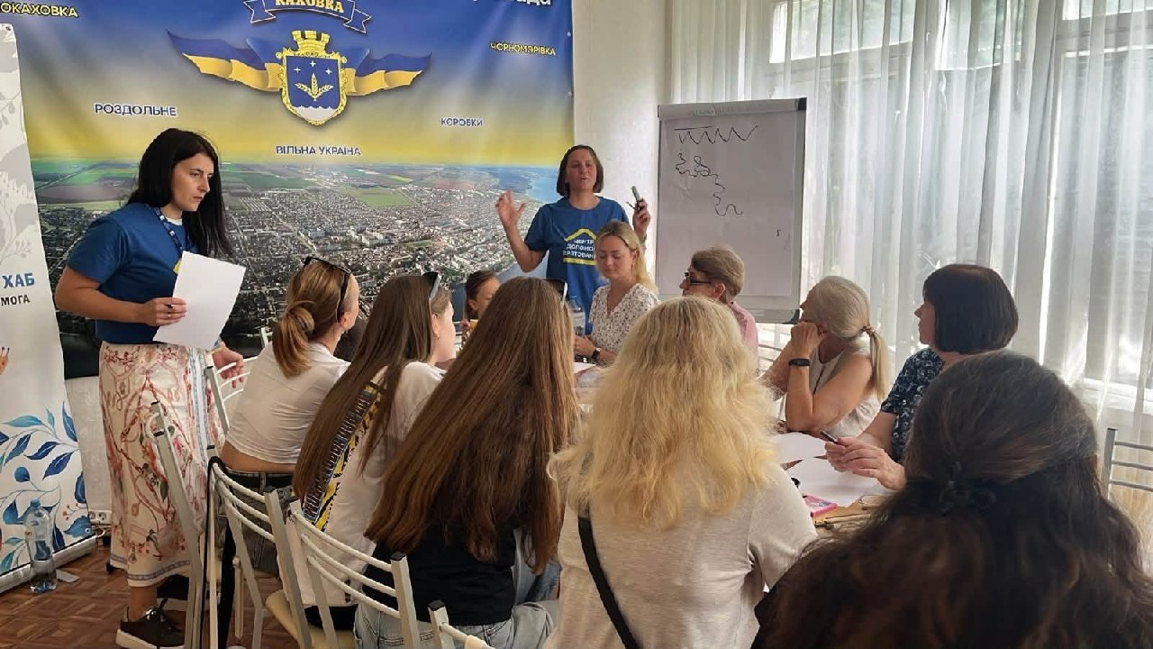 Заняття з нейрографіки для переселенок із Херсонщини в координаційному центрі «Вільні разом» у місті Києві. Фахівчиня у синій фірмовій футболці стоїть біля фліпчарта з маркером у руці та пояснює техніку малювання учасницям, які сидять навколо великого столу, а інша організаторка з аркушем паперу стоїть ліворуч на тлі великого банера Каховки.
