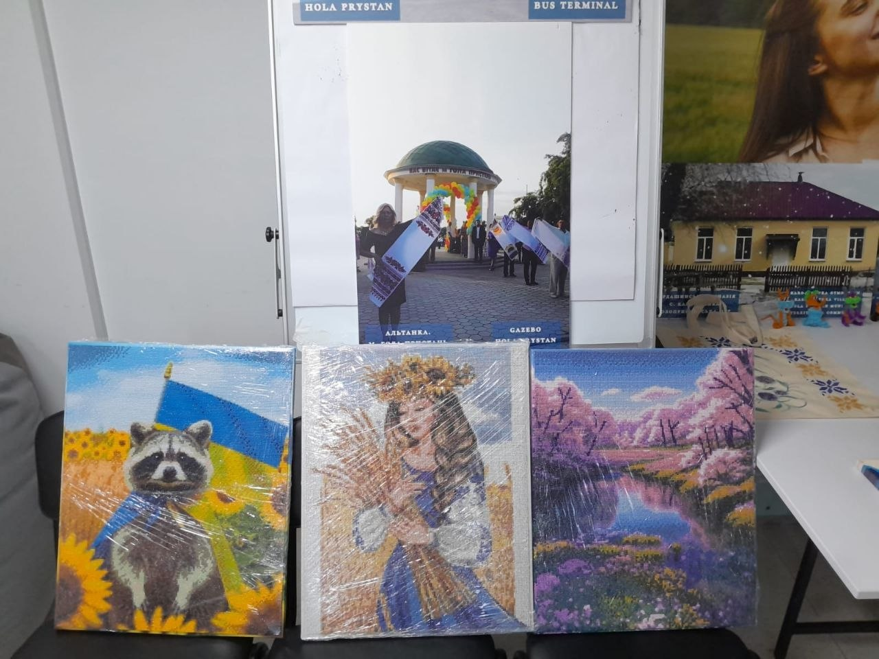 Експозиція фотовиставки та творчих робіт Голопристанської громади в Центрі життєстійкості. На стільцях розставлено три запаковані в плівку картини: патріотичний єнот на тлі соняшників та прапора, дівчина у вінку з колосками пшениці та ліловий пейзаж. На стіні над ними розміщено світлину знаменитої білої альтанки у Голій Пристані до окупації.
