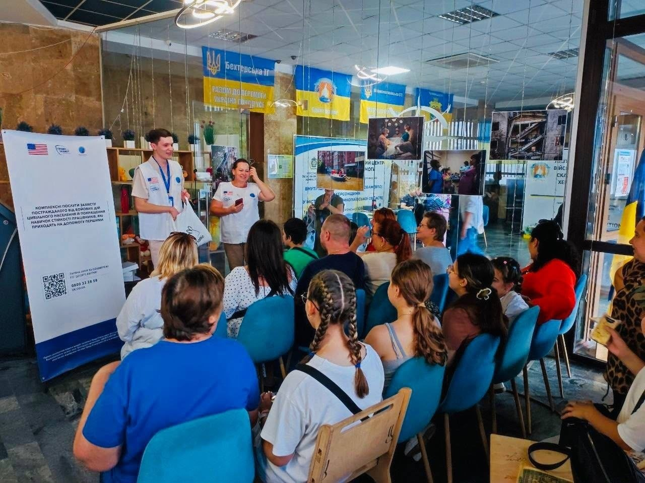 У приміщенні координаційного центру «Вільні разом» у Миколаєві проходить заняття групи психологічної підтримки для переселенців з Херсонщини. На передньому плані спікери від ГО «Десяте квітня» — хлопець та жінка у білих жилетах із логотипами організації та прапорами України і США — виступають перед аудиторією. Слухачі, переважно жінки різного віку, сидять на блакитних стільцях спиною до камери та уважно слухають. Ліворуч стоїть великий інформаційний банер із текстом про комплексну допомогу постраждалим від бойових дій та контактами, а на стінах на фоні розміщені прапори України, символіка організацій та стенди з фотографіями.