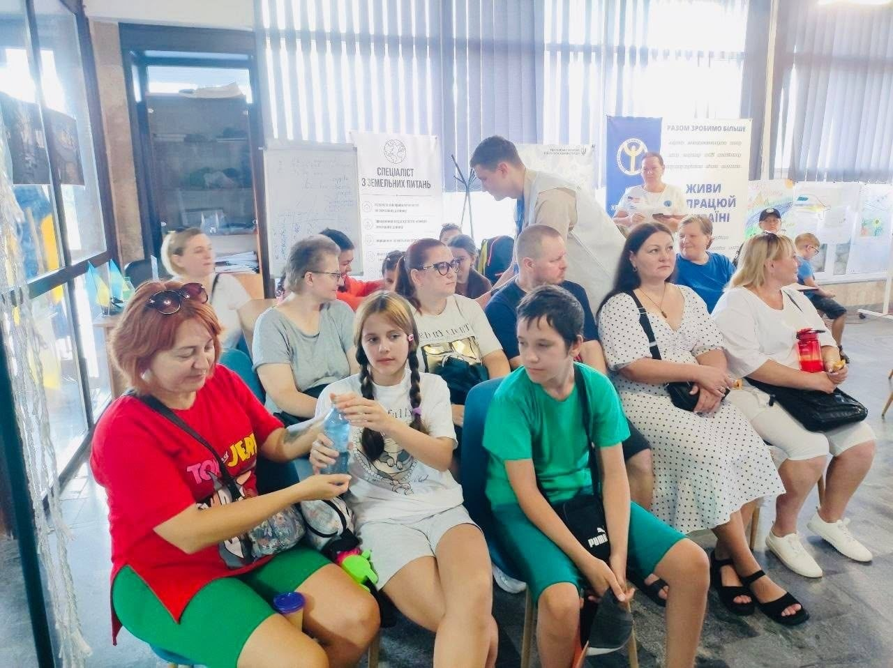 У світлому приміщенні координаційного центру «Вільні разом» на стільцях сидять переселенці з Херсонщини. На передньому плані ліворуч жінка в червоній футболці та зеленавих шортах спілкується з дівчинкою, яка тримає пластикову пляшку води. Поруч із ними сидить хлопчик у зеленій футболці та шортах з логотипом Puma. На задньому плані інші дорослі та діти слухають фахівців. Чоловік-консультант у світлому жилеті ГО «Десяте квітня» нахилився до одного з учасників для індивідуальної бесіди. На фоні видно вертикальні білі жалюзі на вікнах, а також інформаційні стенди, серед яких плакат із написом «Спеціаліст з земельних питань» та синій банер із гаслом «Живи, працюй в Україні».