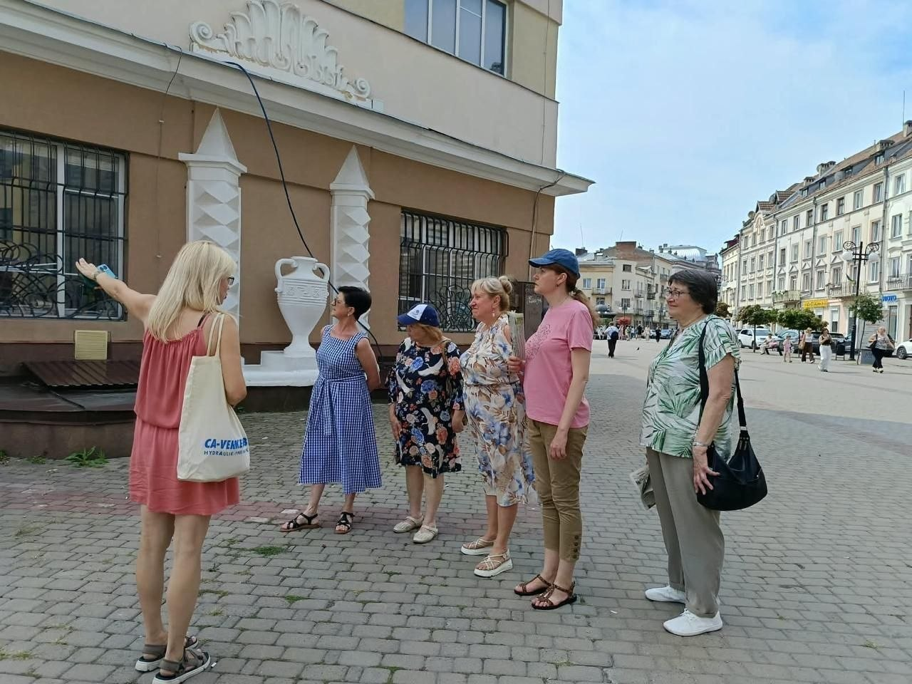 Під час екскурсії «Вулицями Станіславова» в Івано-Франківську група переселенок з Херсонщини разом із гідом зупинилася на бруківці біля історичної споруди. Ліворуч спікерка у рожевій сукні та з тканевою екосумкою через плече показує рукою на фасад будинку, звертаючи увагу на ліпнину у формі мушлі над вікном першого поверху. П'ять учасниць різного віку стоять поруч і уважно розглядають архітектурні елементи, біля яких встановлено велику білу декоративну вазу. Праворуч відкривається перспектива широкої пішохідної вулиці міста з іншими старовинними багатоповерховими кам'яницями під черепичним дахом.