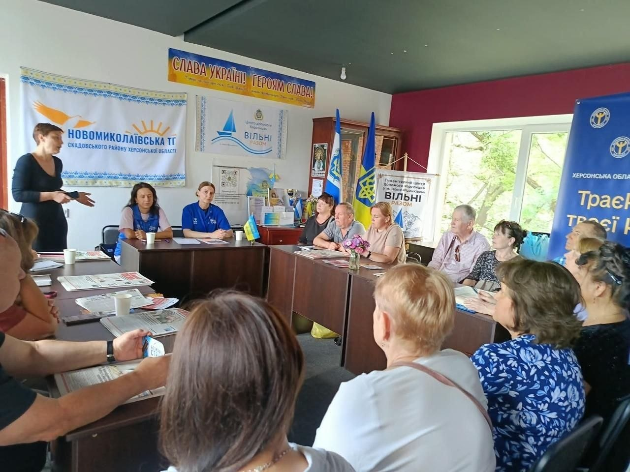 В Івано-Франківську в координаційному центрі «Вільні разом» проходить інформаційна зустріч юристів БФ «Право на захист» для переселенців із Херсонщини. Ліворуч біля столу виступає спікерка в чорній сукні, яка роз'яснює присутнім можливості отримання житла в сільській місцевості. Учасники заходу сидять за темно-коричневими столами й уважно слухають. На стінах кабінету розміщено прапори України, патріотичне гасло «СЛАВА УКРАЇНІ! ГЕРОЯМ СЛАВА!» та інформаційні банери, серед яких стенд «Новомиколаївська ТГ Скадовського району Херсонської області».