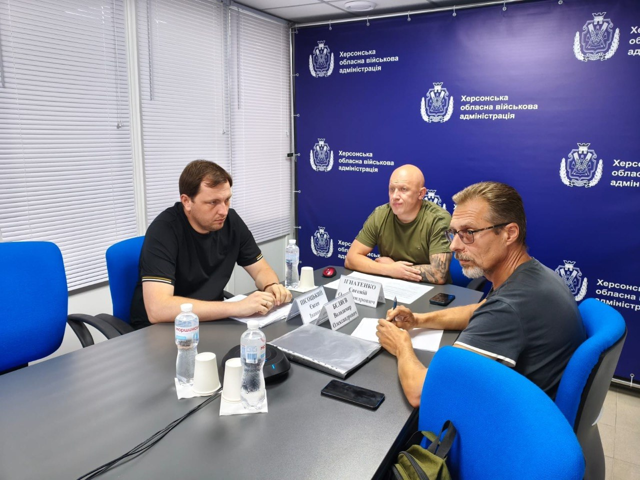 За робочим столом у кабінеті проходять онлайн-засідання щодо евакуації населення керівники Херсонської ОВА. Ліворуч за столом у чорній футболці-поло сидить директор Департаменту з питань цивільного захисту та оборонної роботи ХОВА Євген Пісоцький. Навпроти нього, праворуч в окулярах та темно-сірій футболці, сидить заступник директора - начальник управління планування заходів цивільного захисту та матеріального забезпечення ХОВА Володимир Бєляєв. По центру на задньому плані в мілітарі-футболці кольору хакі сидить заступник начальника Херсонської ОВА Євгеній Ігнатенко. Нарада проходить на тлі великого синього банера з написом «Херсонська обласна військова адміністрація», а на столі перед посадовцями розкладено документи, телефони та встановлено іменні таблички.