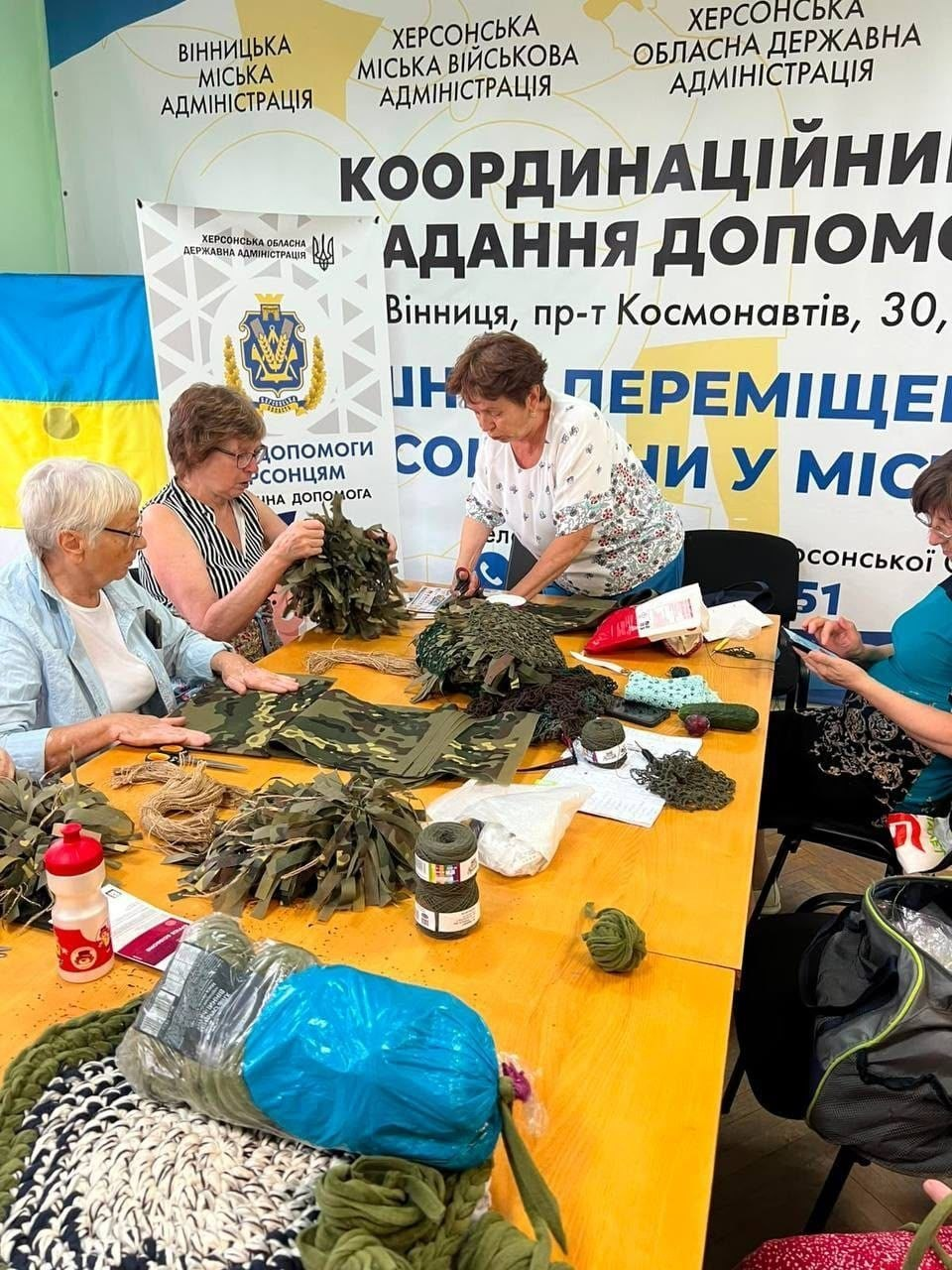 У координаційному центрі «Вільні разом» у Вінниці херсонські переселенки за великим столом виготовляють засоби захисту для ЗСУ — обшивають камуфляжною тканиною плитоноски бронежилетів та плетуть захисні нашоломники. На тлі видно офіційні банери Вінницької міської, а також Херсонської міської та обласної адміністрацій із зазначенням адреси центру.