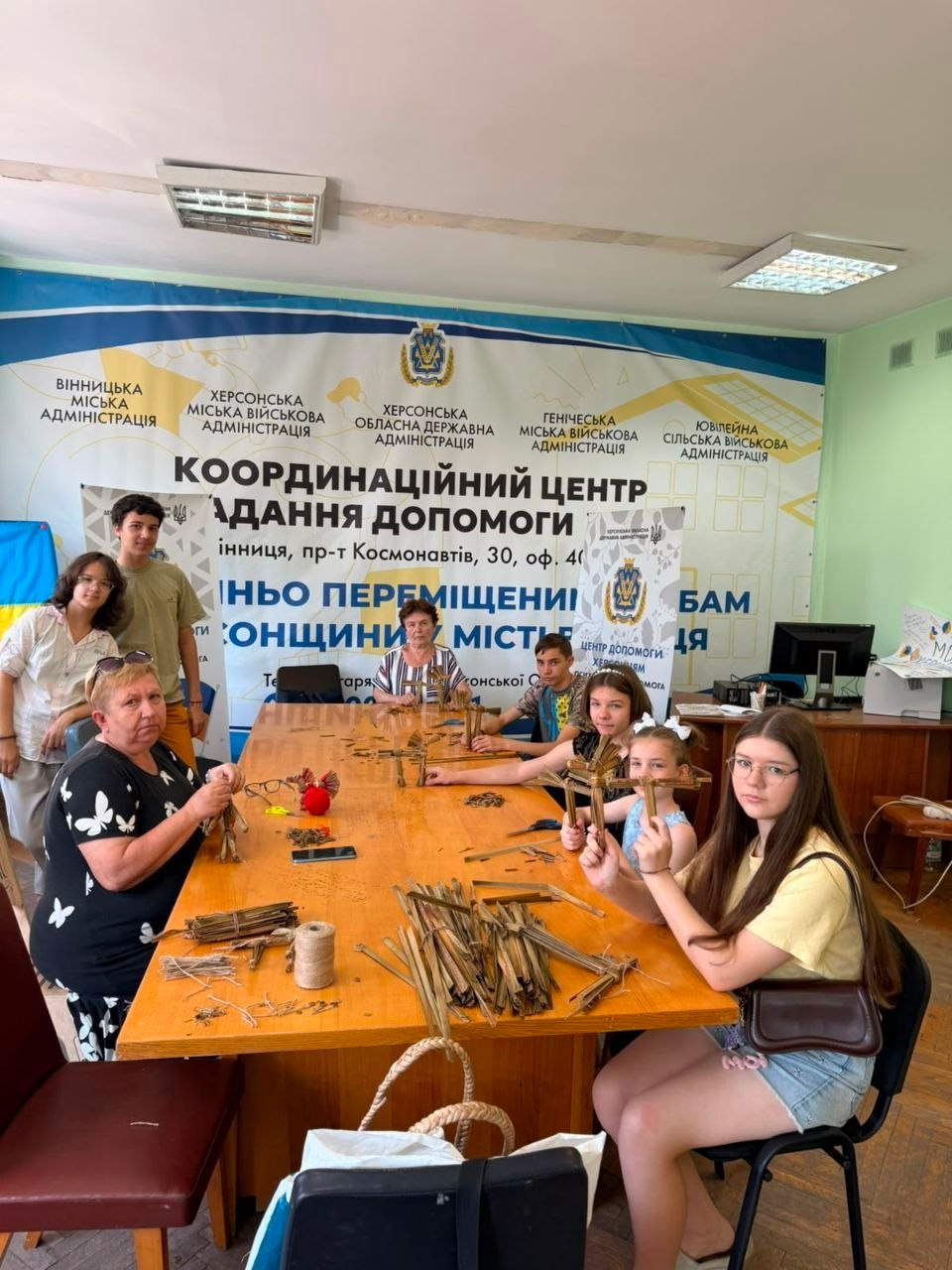 У координаційному центрі «Вільні разом» у Вінниці херсонські переселенці — дорослі, підлітки та діти — сидять за столом і виготовляють традиційні обереги під час майстер-класу зі створення коника з рогозу. На задньому плані видно великий банер центру із символікою Вінницької та Херсонської міських, обласних і сільських адміністрацій.
