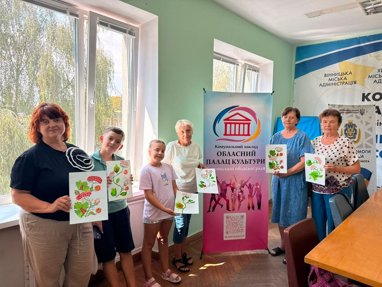 У координаційному центрі «Вільні разом» у Вінниці херсонські переселенці — дорослі жінки та двоє дітей — стоять у світлій кімнаті та показують свої малюнки з гронами калини, створені під час майстер-класу з елементами петриківського розпису. Поруч встановлено яскравий банер Обласного палацу культури Херсонської обласної ради, а позаду видно банери Вінницької та Херсонської міських адміністрацій.