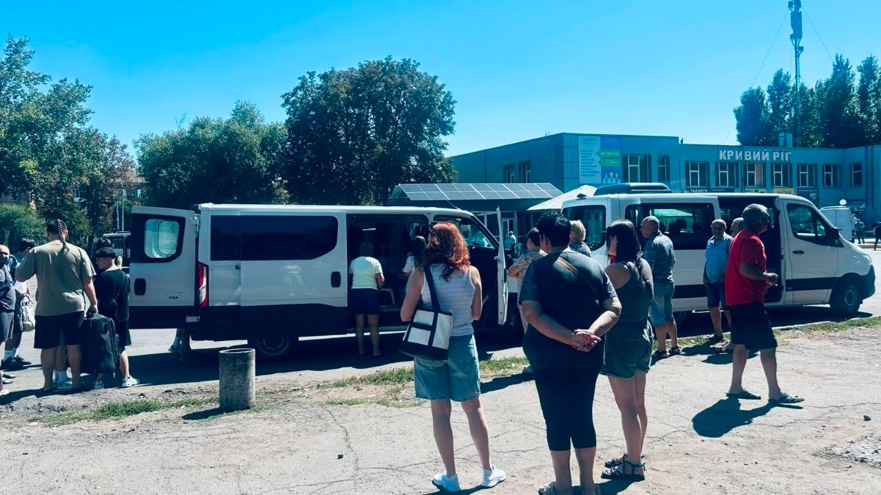Учасники міжнародного обміну з Бериславського академічного ліцею на Херсонщині разом із керівництвом громади стоять біля двох білих мікроавтобусів у Кривому Розі, готуючись до поїздки до Польщі в межах молодіжного проєкту «Місто Надії». На задньому плані видно міську будівлю з написом «КРИВИЙ РІГ», зелені дерева та сонячне небо.