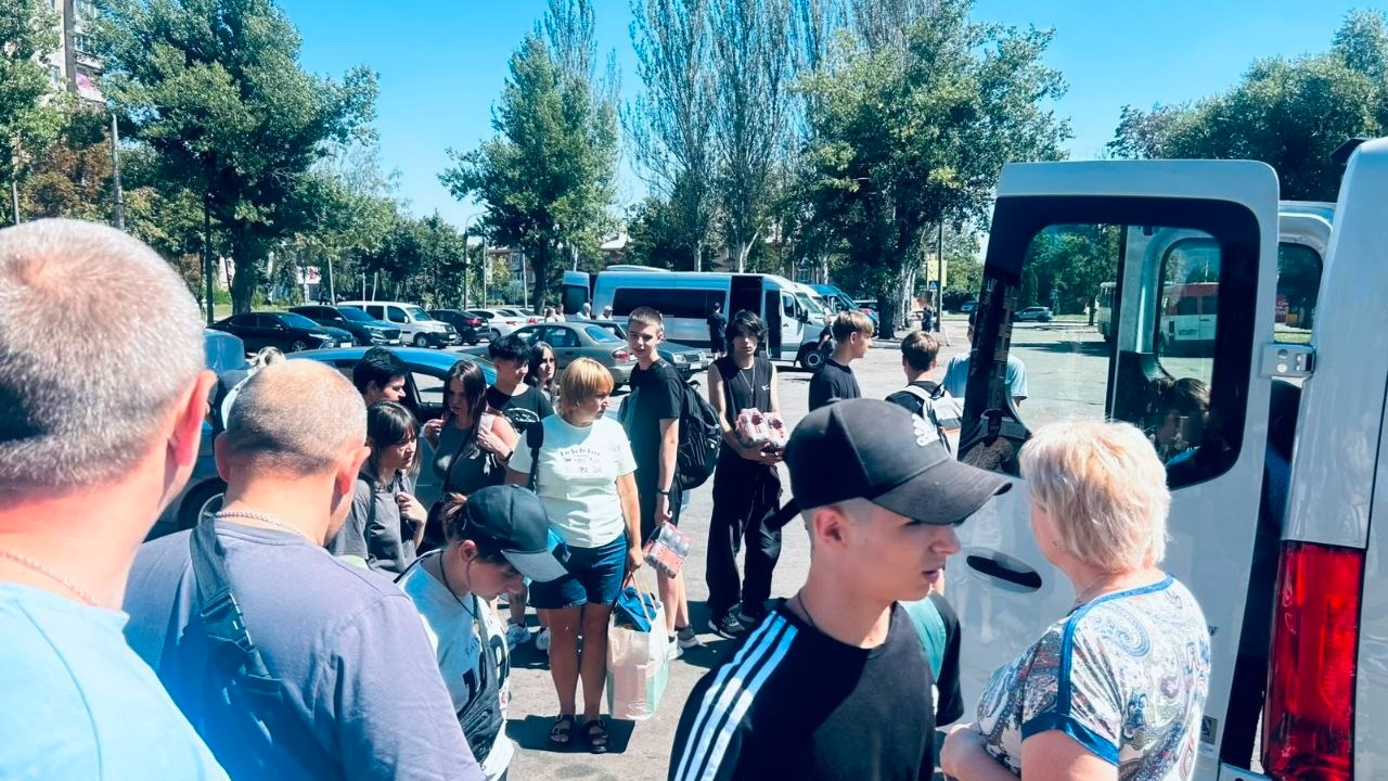 Група підлітків-ліцеїстів із Берислава та дорослих стоїть на вулиці біля відчинених дверей білого мікроавтобуса перед виїздом у міжнародний обмін до Польщі. На передньому плані видно людей у літньому одязі та кепках, а на задньому плані — міську парковку з автомобілями, зелені високі дерева та безхмарне синє небо.
