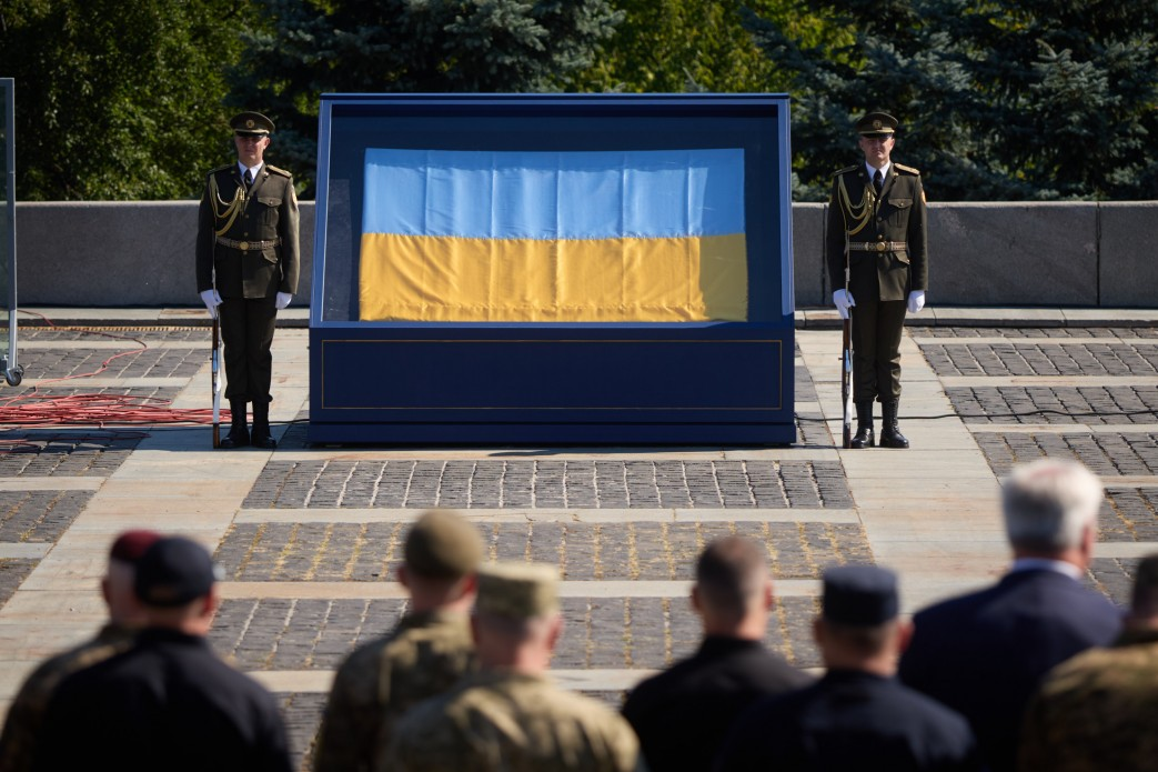 Двоє військовослужбовців почесної варти стоять на варті біля історичного Державного Прапора України, розміщеного у спеціальній захисній вітрині, під час офіційних урочистостей та звернення Президента України Володимира Зеленського з нагоди Дня Державного Прапора. На передньому плані зі спини видно присутніх українських військових та іноземних гостей, які беруть участь у церемонії просто неба.