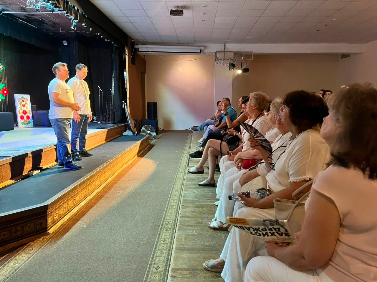 Заступник начальника Херсонської ОДА Володимир Клюцевський у вишиванці виступає на сцені перед глядачами під час показу програми «UA: Музична мапа України» у Херсоні. У глядацькій залі на стільцях сидять мешканці міста, які слухають промову, а жінка на передньому плані тримає буклет із написом «ЗАХИСТ ДЕРЖАВИ».