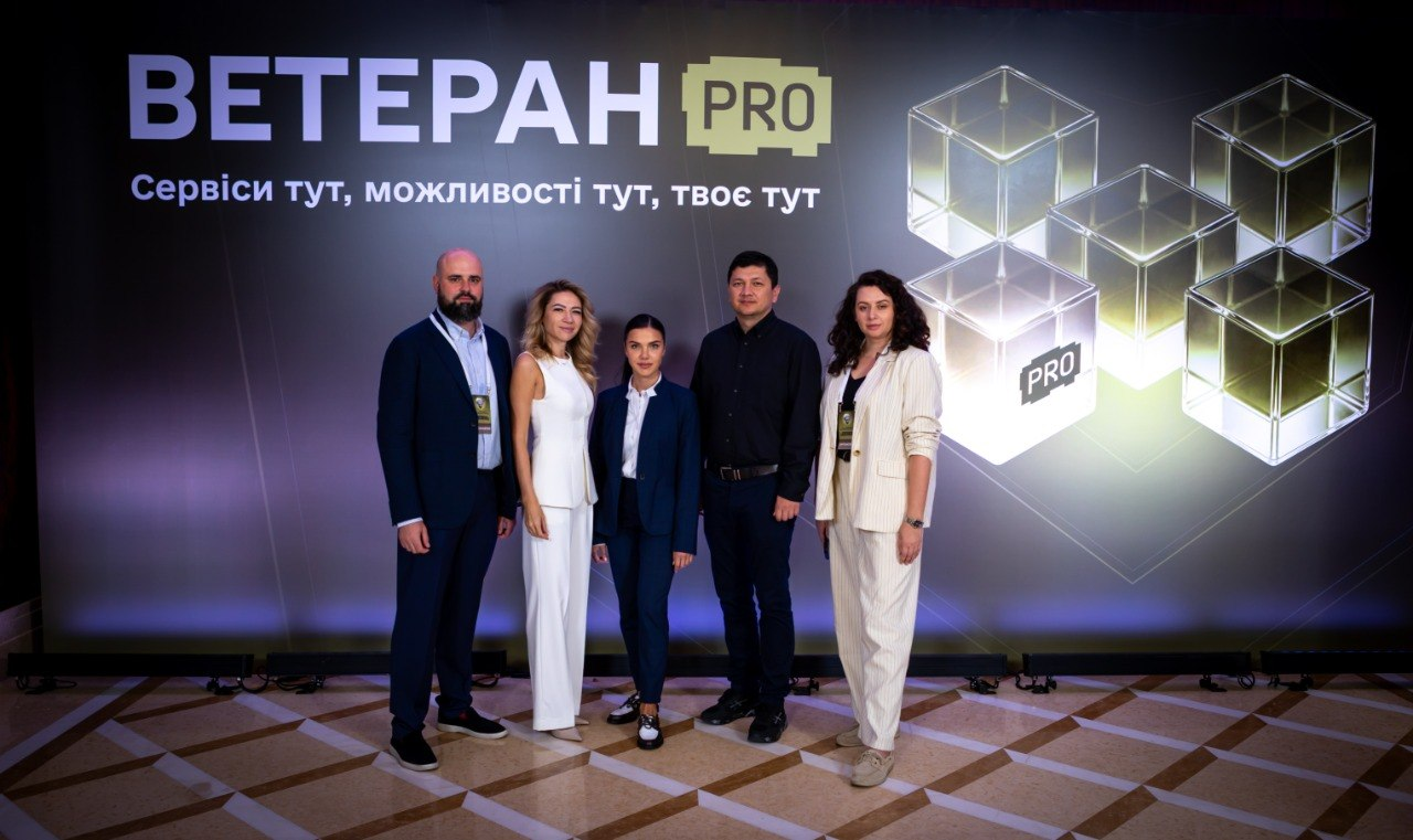 Групове фото п'ятьох учасників Міжнародного ветеранського форуму, серед яких заступниця начальника Херсонської ОВА Ольга Малярчук (друга ліворуч у білому брючному костюмі) та Міністр у справах ветеранів України Віталій Кім (другий праворуч у чорній сорочці), які стоять у повний зріст на тлі офіційного темного банера цифрової платформи «ВЕТЕРАН PRO».