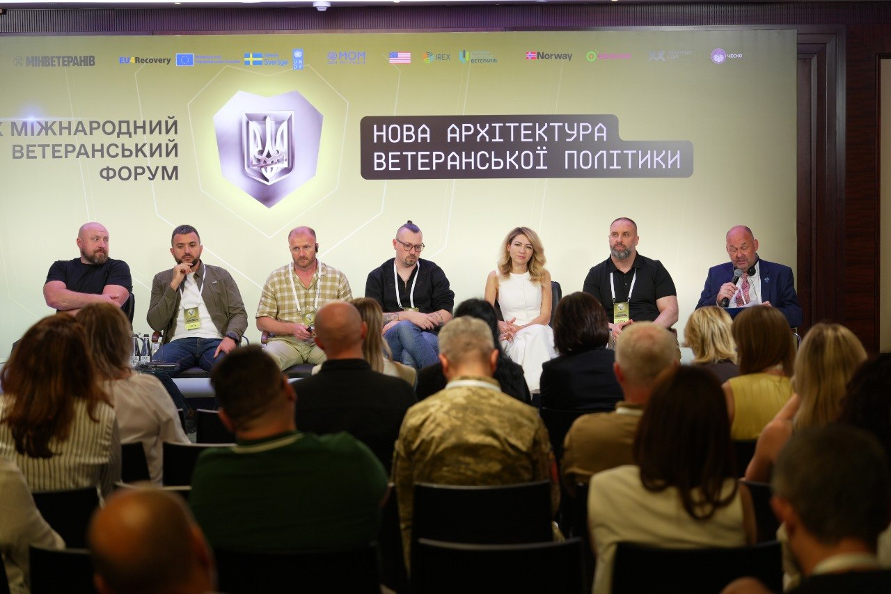 Панельна дискусія на Міжнародному ветеранському форуму «Нова архітектура ветеранської політики». Сім спікерів, серед яких заступниця начальника Херсонської ОВА Ольга Малярчук (у білому костюмі), сидять на сцені та виступають перед аудиторією в залі. На великому світлому банері позаду них розміщено герб України, назву заходу та логотипи міжнародних партнерів.