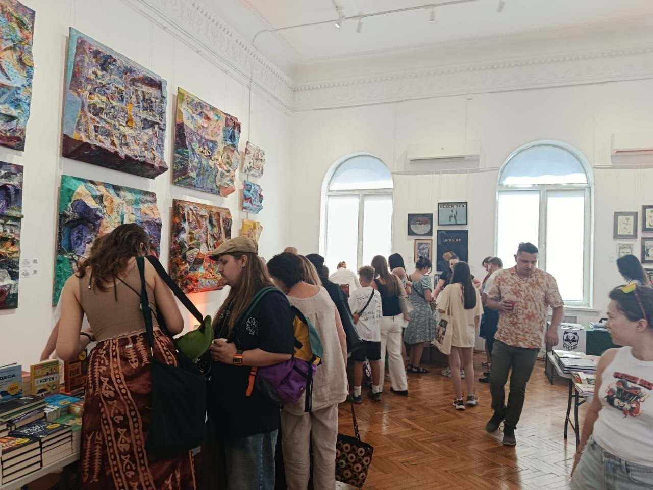 Відвідувачі, серед яких представники координаційного центру «Вільні разом», на книжковому фестивалі «Змістовно» у залі Черкаського художнього музею. Люди розглядають книги на столах, спілкуються та оглядають виставку сучасних об'ємних картин на стінах світлої зали з паркетною підлогою.