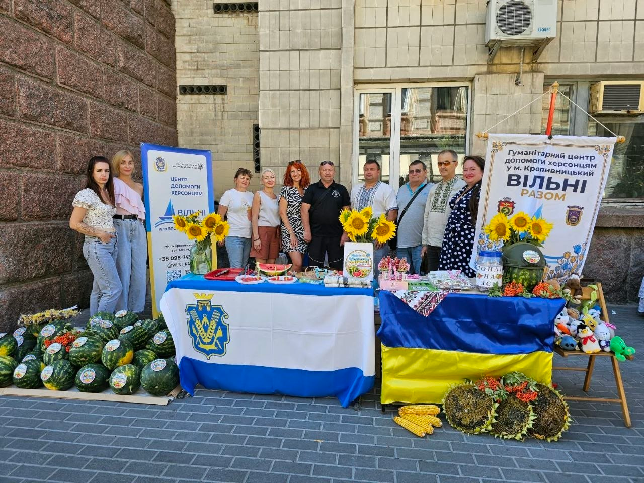 Команда координаційного центру «Вільні разом» на благодійному забігу та аукціоні «WAYPOINT-2026» у Кропивницькому. Волонтери стоять біля презентаційних столів із прапорами України та міста, пропонуючи відвідувачам свіжі херсонські кавуни, кукурудзу, соняшники та патріотичні сувеніри ручної роботи для збору коштів на підтримку ЗСУ.