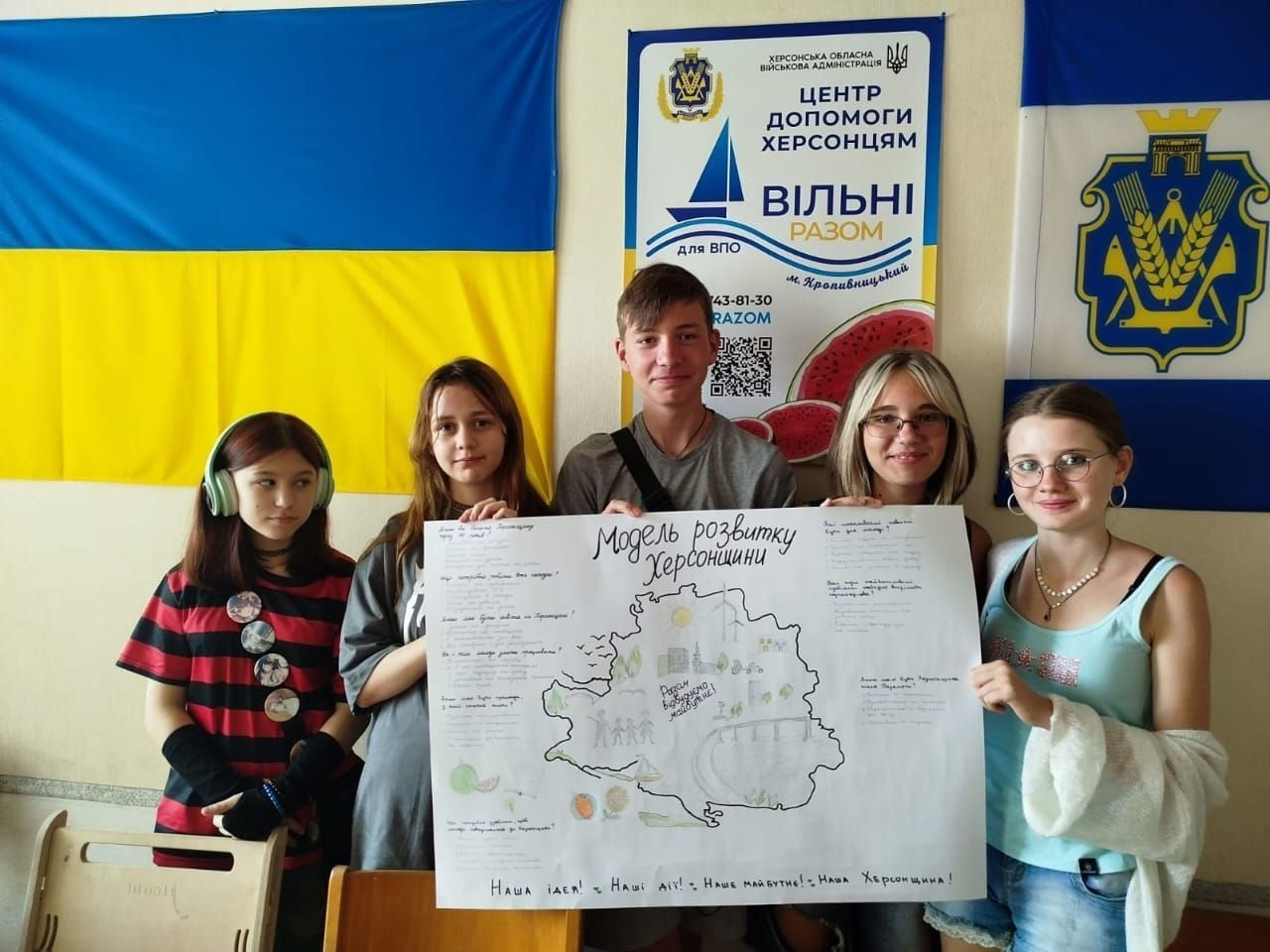 П'ятеро підлітків у кропивницькому координаційному центрі «Вільні разом» тримають великий намальований плакат «Модель розвитку Херсонщини» під час молодіжного флешмобу. Вони стоять на тлі прапора України, прапора Херсонської області та офіційного банера «Центр допомоги херсонцям». На ватмані зображено карту деокупованої області, схематичні вітряки, сонце, людей та патріотичні гасла.
