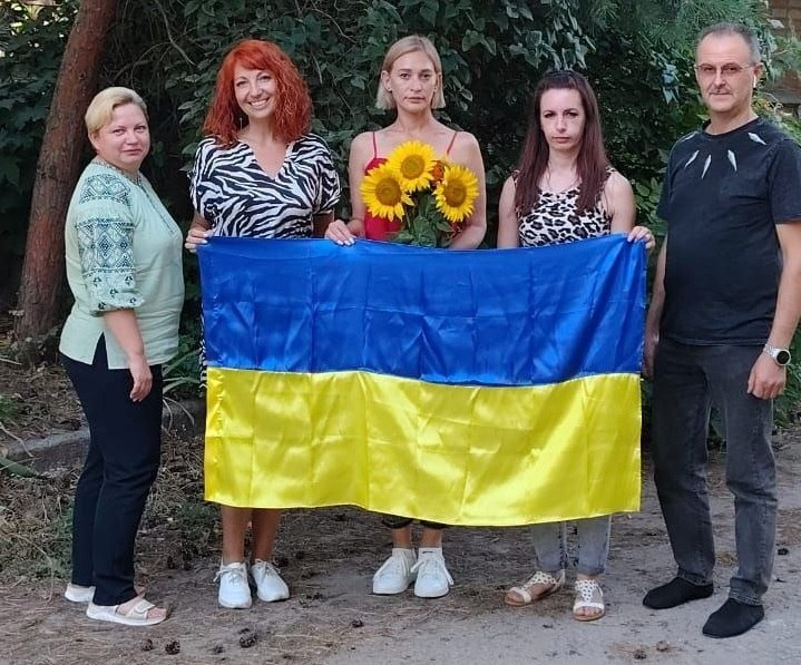 П'ятеро представників та відвідувачів координаційного центру допомоги херсонцям у місті Кропивницький «Вільні разом» разом тримають великий синьо-жовтий прапор України під час урочистостей до Дня Державного Прапора. Учасники стоять у повний зріст на тлі зелених дерев, а жінка у центрі тримає в руках яскравий букет із трьох соняшників.