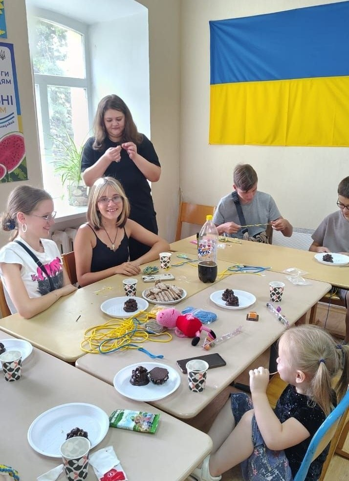 Діти та підлітки з числа ВПО з Херсонщини беруть участь у майстер-класі з нейрографіки та інтерактиві «Кольори емоцій» у просторі «КОМА» координаційного центру «Вільні разом» у місті Кропивницький. Учасники сидять за великим столом, на якому розкладені кольорові шнурки, солодощі та паперові стаканчики, та спілкуються з ментором у чорній футболці. На стіні світлої кімнати висить прапор України та офіційний банер центру.
