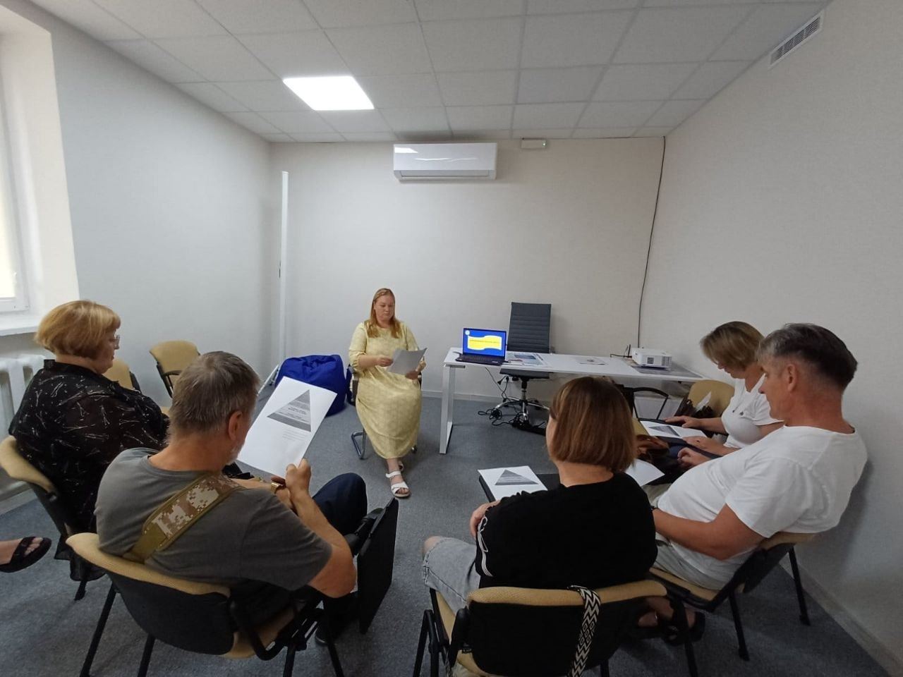 У Координаційному центрі «Вільні разом» у Житомирі відбувся вебінар «Власна справа» від Херсонського обласного центру зайнятості для внутрішньо переміщених осіб з Херсонщини. У світлому офісному кабінеті жінка-модератор у жовтій сукні провела зустріч для п'ятьох учасників, які розмістилися півколом на стільцях. Слухачі активно опрацьовували друковані інформаційні матеріали, зокрема графіки-піраміди, а на робочому столі поруч із проєктором було розгорнуто ноутбук із символічним зображенням прапора України на екрані.