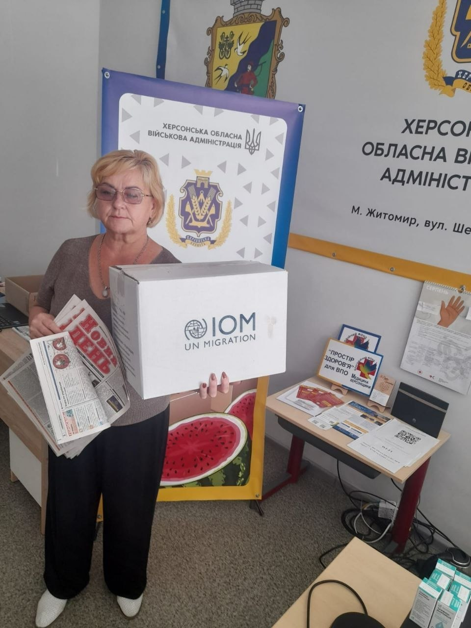 У Координаційному центрі «Вільні разом» у Житомирі триває системна підтримка внутрішньо переміщених осіб з Херсонщини. На фото жінка отримує гуманітарну допомогу — велику білу коробку від Міжнародної організації з міграції (IOM UN MIGRATION) та свіжі випуски газети «Новий день» на тлі символіки та банерів Херсонської обласної військової адміністрації.