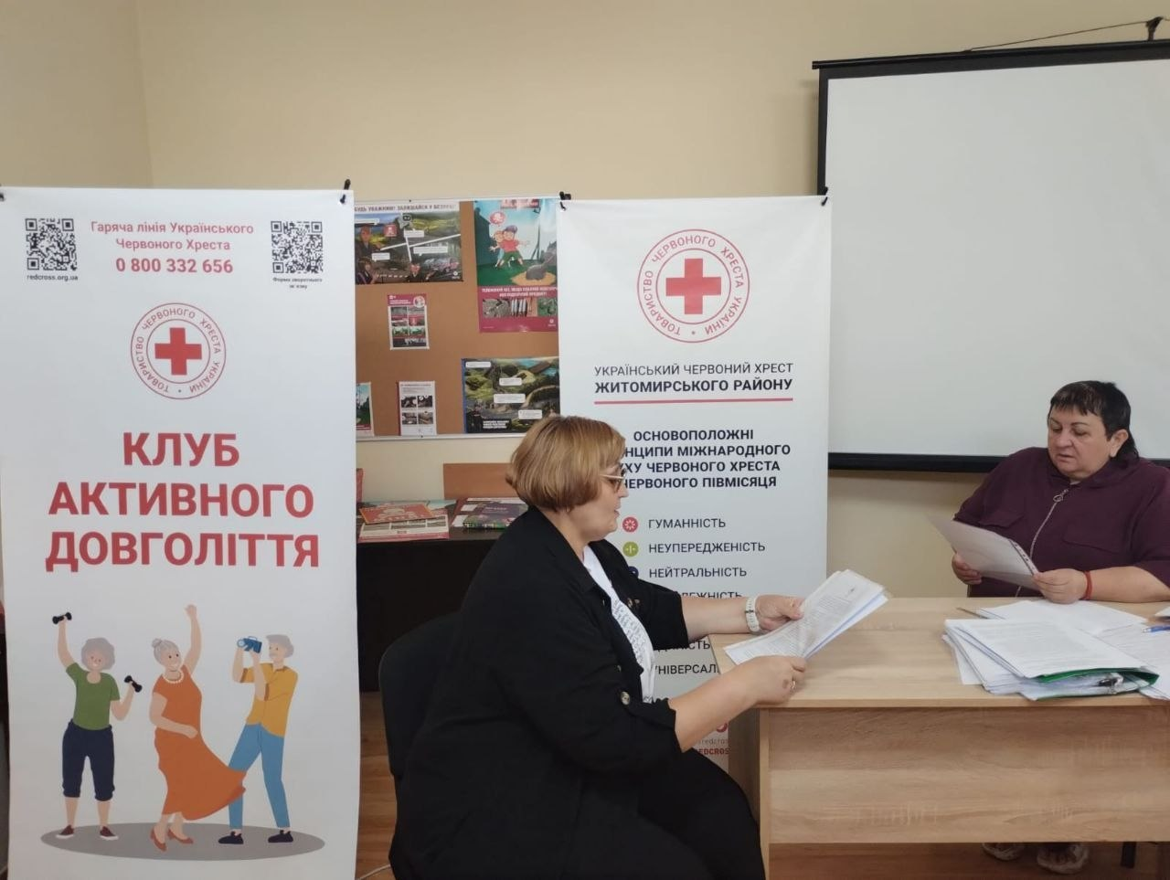 У Координаційному центрі «Вільні разом» у Житомирі в межах системної підтримки внутрішньо переміщених осіб з Херсонщини відбулася зустріч із головою ГО «Міськрайонна організація Товариства Червоного Хреста України» Раїсою Черемісіною. На фото дві жінки працюють із документами за робочим столом на тлі інформаційних банерів «Клуб активного довголіття» та «Український Червоний Хрест Житомирського району».